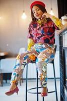 Enthusiastic african american woman in trendy coloured outfit with red beret chilling in cozy cafe, sitting on bar counter. photo