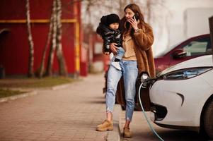 Young mother with child charging electro car at the electric gas station and speak on mobile phone. photo