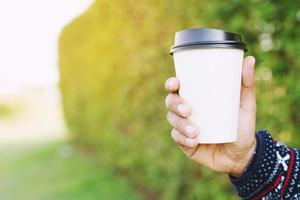 young hand holding paper cup of take away drinking coffee on natural morning sunlight. space Place for your text or logo.