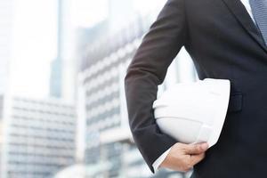 Close up front view of engineering business man suit contractor construction worker holding safety white helmet for the safety of the work operation. outdoor of building background. photo