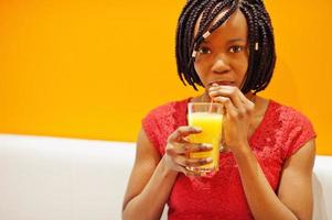 African woman sitting on cafe against orange wall and drink pineapple juice in hands. photo