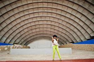 Amazing african american model woman in green pants and black hat posed outdoor against arena hall. photo