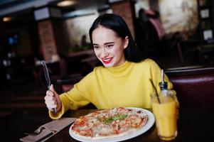 Funny brunette girl in yellow sweater eating pizza at restaurant. photo