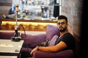 Stylish beard arabian man in glasses and black t-shirt smoking hookah indoor bar. Arab model having rest. photo