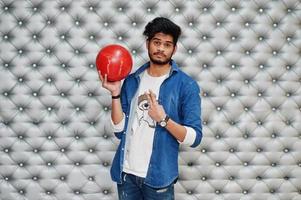 Stylish asian man in jeans shirt standing with bowling ball at hand against silver wall background and show two fingers. photo