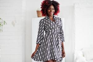 Fashionable tall african american model woman with red afro hair in dress posed at white room against vintage wardrobe. photo