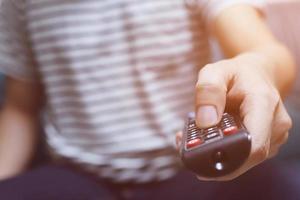 close up Television remote control in casual man hands pointing to tv set and turning it on or off. select channel watching tv on his sofa at home in the living room relax. photo