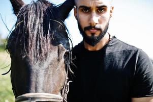 Hombre árabe de barba alta vestido de negro con caballo árabe. foto