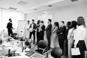 Mixed race business coach presenting report standing near whiteboard pointing on sales statistic shown on diagram and chart teach diverse company members gathered together in conference room. photo