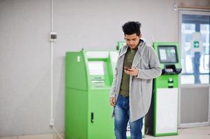 joven asiático con estilo con teléfono móvil contra la fila de cajero automático verde. foto