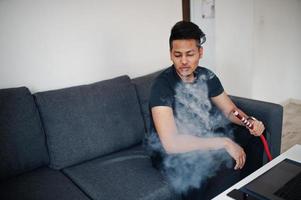 Handsome and fashionable indian man in black sitting at room, smoking hookah photo