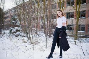 modelo morena de piernas largas de moda en una larga capa negra posada al aire libre en el día de invierno contra una fábrica abandonada. foto