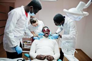 Multiracial dentist doctors team. African american man patient at UV protective glasses. His teeth treated with the help of a dental UV curing light lamp and a dental mirror. photo
