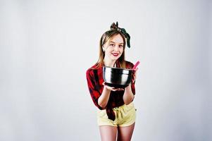 Young funny housewife in checkered shirt and yellow shorts pin up style with saucepan and kitchen spoon isolated on white background. photo