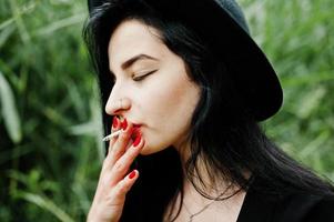 Sensual smoker girl all in black, red lips and hat. Goth dramatic woman smoking on common reed. photo