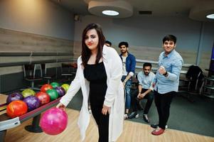 South asian woman standing at bowling alley with ball on hands. Girl is preparing for a throw. Friends support her loudly. photo