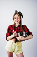 Young funny housewife in checkered shirt and yellow shorts pin up style with saucepan and kitchen spoon isolated on white background. photo