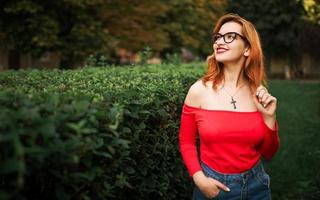 atractiva mujer pelirroja con anteojos, vestida con blusa roja y falda de jeans posando en el parque verde. foto