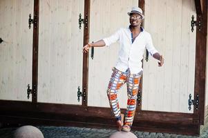 Stylish african american man in white shirt and colored pants with hat and glasses posed outdoor. Black fashionable model boy. photo