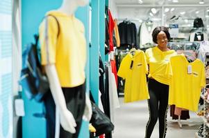 Afican american women in tracksuits shopping at sportswear mall against shelves. She choose yellow t-shirt. Sport store theme. photo