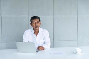 Work from home and social distancing concept. Asian business old man  working and drinking coffee at co-working space. Aging man using laptop meeting online. photo