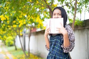 una niña sostiene un libro y está parada debajo de un hermoso árbol. foto