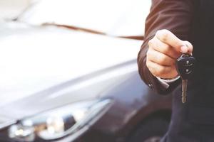 hombre de negocios mano sujetando las llaves del coche delante con un coche nuevo en el fondo. estacionamiento en frente de la casa. concepto de transporte. deje espacio de copia para escribir mensajes de texto. foto