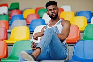 Handsome african american man at jeans overalls posed on colored chairs at stadium. Fashionable black man portrait. photo