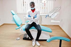 African american male doctor in mask with crossed arms sitting at dentist chair in dental clinic. photo