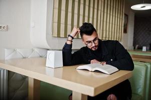 Arab man wear on black jeans jacket and eyeglasses sitting in cafe, read book. Stylish and fashionable arabian model guy. photo