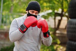 retrato deportivo boxeador árabe con mascarilla médica negra boxeando al aire libre durante la cuarentena del coronavirus. foto