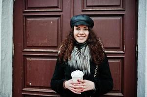 Curly mexican girl in leather cap and plastic cup of coffee at hand walking at streets of city. photo