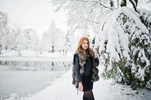 Red haired girl in fur coat walking at winter snowy park. photo