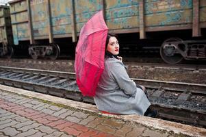 Brunette girl in gray coat with red umbrella in railway station. photo