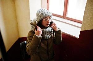 Portrait of brunette girl in gray scarf and hat, glasses near window. photo