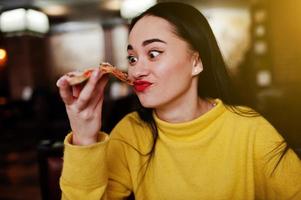 Funny brunette girl in yellow sweater eating pizza at restaurant. photo