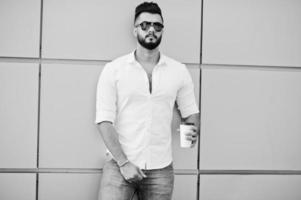 Stylish tall arabian man model in white shirt, jeans and sunglasses posed at street of city. Beard attractive arab guy with cup of coffee against golden wall. photo