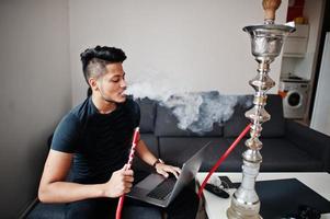 Handsome and fashionable indian man in black sitting at room, smoking hookah and working on laptop. photo