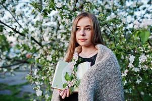 Young brunette girl on plaid against spring blossom tree and holding book at hands. photo