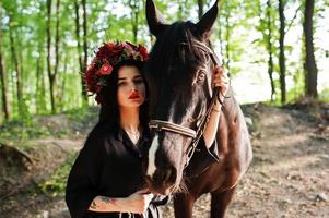 Mystical girl in wreath wear in black with horse in wood. photo