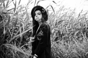 Sensual smoker girl all in black, red lips and hat. Goth dramatic woman smoking on common reed. Black and white portrait. photo