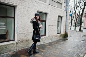 Curly mexican girl in leather cap and plastic cup of coffee at hand walking at streets of city. photo