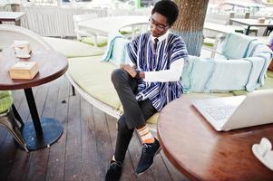 African man in traditional clothes and glasses sitting behind laptop at outdoor caffe. photo