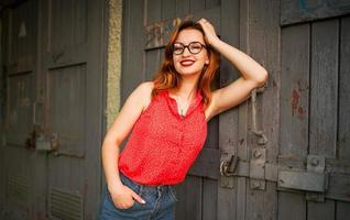 atractiva mujer pelirroja con anteojos, vestida con blusa roja y falda de jeans posando. foto