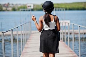 Back of stylish african american model in glasses hat, jeans jacket and black skirt posed outdoor on pier against lake. photo