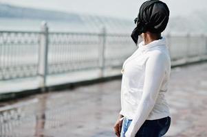 chica musulmana africana con gafas de sol, hiyab negro, sudadera blanca y jeans posados al aire libre contra la fuente. foto