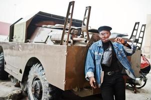 African american man in jeans jacket, beret and eyeglasses, smoking cigar and posed against btr military armored vehicle. photo