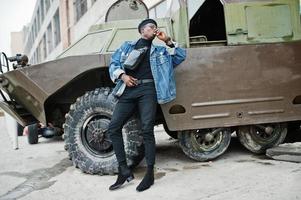 African american man in jeans jacket, beret and eyeglasses, smoking cigar and posed against btr military armored vehicle. photo