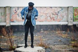 hombre afroamericano con chaqueta de jeans, boina y anteojos contra la pared de graffiti en un techo abandonado. foto