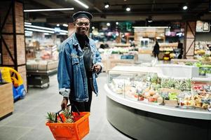 Elegante hombre afroamericano casual con chaqueta de jeans y boina negra sosteniendo una canasta con piñas en la sección orgánica de frutas del supermercado. foto